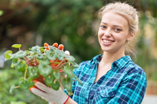 A young woman with blonde hair tied back, smiling while holding a small potted flowering plant with green and reddish leaves, standing outdoors in a garden setting. The background features blurred greenery, including trees and shrubs, with natural daylight illuminating the scene. She is wearing a blue and black checked shirt and white gardening gloves, indicating involvement in gardening or outdoor maintenance activities. The environment appears to be a well-maintained residential garden or landscaped outdoor space, with a mix of trees and plants that could be part of a typical Edmonton home garden, reflecting local gardening practices and landscape features associated with the area. The scene supports gardening services such as planting and garden care, highlighting an active engagement with outdoor landscaping activities.