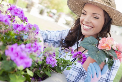 A smiling woman wearing a straw gardening hat, light blue gloves, and a blue checkered shirt is tending to flowering plants in a well-maintained garden during daytime. In front of her are vibrant purple and pink flowers, with green foliage and leaves of different shades. The background features neatly mowed grass, a paved pathway, and some trees, suggesting a peaceful outdoor space in Edmonton. The scene reflects outdoor gardening activity, with the woman carefully inspecting or arranging the plants, supported by professional landscaping and garden maintenance services that enhance the appearance and health of outdoor gardens in the local area.