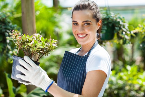 A smiling woman with dark hair tied back, wearing a white t-shirt, a blue striped apron, and white gardening gloves, is standing outdoors in a well-maintained garden. She holds a small potted shrub with green and reddish leaves, with lush green foliage and flowering plants in the background. The garden features a wooden tree stake, dense greenery, and well-kept flower beds, indicating healthy plant growth. Bright natural daylight illuminates the scene, suggesting clear weather. The setting appears to be a landscaped yard, possibly part of a residential garden in Edmonton, with a focus on gardening and outdoor maintenance, reflecting professional gardening services provided by Landscapers Edmonton.