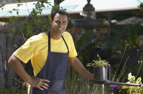 A man with medium skin tone, short dark hair, and a bright smile stands in a well-maintained backyard garden during daytime. He is wearing a yellow T-shirt and a dark grey apron, and is holding a small black pot containing a young plant with thin green leaves. The garden features a lush green lawn with dense, spiky grass in the foreground, bordered by a gravel pathway. In the background, there are various shrubs, small trees, and flowering plants, along with a wooden fence and a decorative garden structure partially visible. The outdoor space appears well-kept with natural light illuminating the scene, highlighting the vibrant greenery and the man's cheerful expression, demonstrating gardening activities in a suburban Edmonton garden, possibly part of a garden renovation project by Landscapers Edmonton.