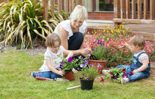 A woman with shoulder-length blonde hair is sitting on a well-maintained grassy lawn in a backyard, engaging with two young children who are planting and tending to colorful flowers in small pots. The children, both with light brown hair and wearing casual summer clothing, are focused on their gardening tasks, with one child holding a purple-flowered plant and the other carefully watering a potted plant with a small watering can. In the background, there is a wooden deck with some potted plants and a cluster of tall, leafy ornamental grasses along the edge of the garden. The scene is captured in natural daylight with a clear weather, highlighting a neatly landscaped outdoor space designed for gardening and family activities, which complements the services offered by Landscapers Edmonton for gardening and outdoor maintenance in the local Edmonton area, near postcode T5A.