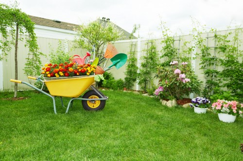 A neatly maintained backyard garden featuring a lush, green lawn with uniform grass density, bordered by a white painted fence and a variety of flowering plants and shrubs, including pink and purple blooms in white pots, arranged along the fence line. The garden has a small tree with light green foliage positioned near the corner, providing partial shade. An outdoor yellow wheelbarrow filled with brightly coloured flowers, gardening tools, and watering cans is situated on the lawn, indicating recent garden maintenance. The background shows the rooftops of neighbouring houses, and the overall scene suggests a calm, well-kept outdoor space suitable for gardening activities and outdoor relaxation, with natural lighting implying a partly cloudy day. This environment reflects typical garden landscaping and lawn care features seen in Edmonton and other UK gardens, aligning with professional gardening and sustainable outdoor maintenance practices by Landscapers Edmonton.