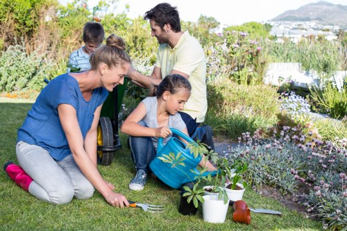 A family of four engaging in gardening activities in a well-maintained outdoor garden space, featuring a lush, green lawn with a variety of flowering plants and shrubs along the borders. The foreground shows a woman kneeling on the grass, planting or tending to a small shrub, while a young girl holds a blue watering can nearby, ready to water the plants. In the background, a man and a boy are sitting on the grass, with the man smiling and supervising the youngsters. The garden includes decorative garden tools, a white watering can, and small pots, all set against a backdrop of trees and flowering borders, with natural daylight and a clear sky, indicating a pleasant and sunny weather typical of outdoor gardening in Edmonton. The scene exemplifies landscape maintenance and planting activities, aligned with professional gardening services by Landscapers Edmonton, focused on creating healthy, vibrant outdoor environments in local residential yards.