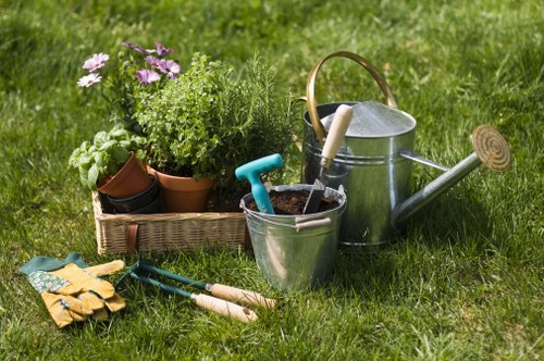 A neatly maintained front or backyard garden features a well-kept green lawn with a variety of gardening tools and equipment arranged on it. In the foreground, there are gardening gloves, small hand trowels, and a pair of pruning shears laid out on the grass. To the side, a metal watering can with a wooden handle and a funnel is positioned next to a small galvanized bucket containing soil. Behind these items, a rectangular wicker basket holds a collection of potted plants, including flowering plants and lush green foliage, with some pots made of terracotta and others plastic. The background reveals a dense line of hedges and shrubs, creating a natural border for the garden, while the overall environment appears to be outdoors on a bright, sunny day, with natural light illuminating the scene and casting soft shadows. The setting suggests an outdoor space suitable for gardening and landscaping work, aligned with services offered by Landscapers Edmonton for maintaining and enhancing garden landscapes in the local area near Edmonton, Alberta.