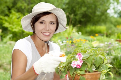A woman wearing a white sun hat, gardening gloves, and a light-colored short-sleeved shirt is smiling while tending to a potted flowering plant with pink and white blossoms. She is in an outdoor garden setting with lush green foliage, including trees and various flowering plants, visible in the background. The garden features a mix of grass, flower beds, and leafy shrubs, with some soil and paving stones also visible. The scene suggests a well-maintained outdoor space suitable for gardening services such as planting, pruning, or lawn care, with natural daylight illuminating the vibrant colors of the flowers and greenery, indicative of a mild weather day in Edmonton, which aligns with local garden environments and professional landscaping standards.