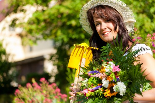 A woman with shoulder-length brown hair and a wide-brimmed straw hat is standing outdoors in a garden, holding a bright yellow gardening tool in her right hand and a large, colorful bouquet of mixed flowers and green foliage in her left arm. Behind her, the garden features a lush green lawn with well-maintained grass, bordered by a variety of flowering plants and shrubs. In the background, there are mature trees with leafy branches providing dappled sunlight, and a blurred house or building structure suggests a residential outdoor space typical of a landscaped front or backyard area in Edmonton. The scene appears to be bright and sunny, with natural light highlighting the vibrant colors of the flowers and the greenery, indicating a warm and pleasant day suitable for gardening and outdoor maintenance activities, reflecting professional gardening service environments in the local area.
