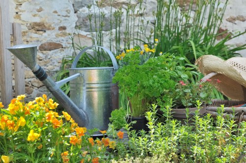 The image depicts a lush garden corner with vibrant yellow and orange flowering plants in the foreground, accompanied by green foliage and small shrubs. Behind the plants, there is a weathered stone wall that adds a rustic backdrop, and a collection of gardening tools and accessories is arranged on the ground. These include a silver metal watering can, a small hand trowel with a black handle, a round planter containing a leafy green herb plant, and a wide-brimmed straw hat resting on a container. Tall, grassy plants are visible in the background, enhancing the natural outdoor environment typical of a landscaped yard. The scene suggests a well-maintained garden area, ideal for gardening services focused on plant care and outdoor space management in the Edmonton or nearby regions. The lighting indicates a bright, possibly sunny day, highlighting the natural textures and colours of the garden elements and creating a warm, inviting outdoor atmosphere.