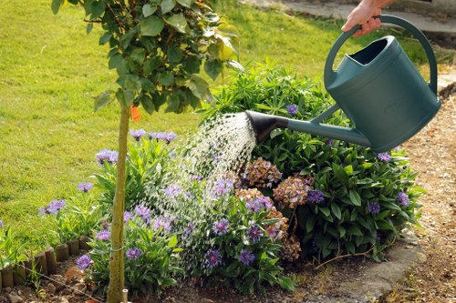 A person watering a garden bed in a residential outdoor space with a green watering can, surrounded by a variety of flowering plants with purple and brown blossoms, and lush green foliage. The garden features a curved border made of wooden edging, with a well-maintained grassy lawn in the background. There is a small tree with green leaves on the left side of the image, and the garden bed appears healthy and vibrant, indicating regular lawn and gardening care. The ground surface around the flower bed is a mixture of soil and paving, with natural sunlight illuminating the scene, suggesting it is a clear day. This setting exemplifies typical gardening practices that Landscapers Edmonton might provide, highlighting landscape maintenance and plant care services in Edmonton, UK, with an emphasis on lush lawns and flourishing flower beds.