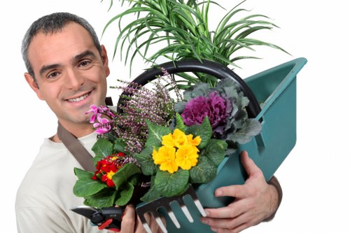 A professional gardener holding a teal plastic watering can filled with a variety of fresh plants and flowers, including purple, yellow, and red blooms, along with lush green foliage. The background features a well-maintained outdoor garden space with a neatly trimmed lawn, small shrubs, and a backdrop of wooden decking or paving stones. The garden appears to be in good condition with a natural, inviting atmosphere, consistent with gardening and landscape maintenance services in Edmonton. The scene is captured outdoors during daylight, with natural light highlighting the vibrant colours and textures of the plants. This image emphasizes outdoor garden care, planting, and maintenance work typical of professional landscaping services available locally.