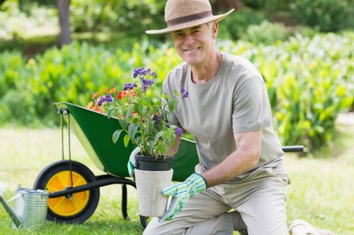 A middle-aged man wearing a straw hat, grey t-shirt, and gardening gloves is kneeling on a well-maintained lawn in a garden, holding a potted plant with blooming purple and red flowers. Behind him, there is a vibrant green hedge and a lush background of trees, indicating a healthy, landscaped outdoor space in Edmonton. To his right, a yellow-wheeled garden wheelbarrow is positioned, filled with garden tools and supplies, suggesting ongoing garden maintenance or planting activities. The garden features a mix of grasses, flower beds, and natural soil areas, with a tidy and organized appearance. Bright daylight illuminates the scene, highlighting the fresh, natural tones of the foliage and flowers, consistent with seasonal conditions in Alberta. This image reflects professional outdoor gardening and landscape enhancement services provided by Landscapers Edmonton, focused on enhancing garden aesthetics and health in local residential environments.