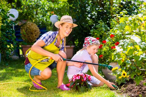 In a lush backyard garden, a woman and a young girl are engaged in gardening activities. The woman, wearing a straw hat, yellow apron, and casual denim shorts, is kneeling on the grass and holding a garden hose, smiling towards the camera. The girl, dressed in a pink hat, white shirt, and pink shoes, is also kneeling and carefully planting or tending to colorful flowers in a flower bed bordered by soil and vibrant blooms, including yellow and red flowers. The garden features a well-maintained grassy lawn with a variety of shrubs, trees, and a dense green hedge in the background, providing a private and inviting outdoor environment. String lights hang overhead, and gardening tools and pots are visible, indicating ongoing planting and lawn care. The scene captures a bright, sunny day in an outdoor space typical of residential gardening services in Edmonton, with natural lighting highlighting the lush foliage and cultivated garden area. This image aligns with professional outdoor landscaping and planting activities offered by Landscapers Edmonton, emphasizing healthy, vibrant gardens and well-kept green spaces in a suburban setting near Edmonton's postcode area.