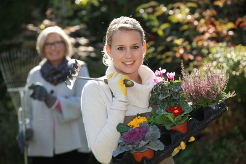A young woman with blonde hair, smiling, holds a tray of colorful flowering plants, including purple, yellow, and pink blooms, in a garden setting during daylight. She is dressed in a light-colored sweater and gardening gloves, with a background of lush greenery and trees with autumnal foliage. Behind her, an older woman with glasses and a scarf is smiling and holding a garden rake, also dressed in light outdoor clothing, suggesting they are engaged in gardening or landscaping activities in a well-maintained, landscaped backyard. The scene features vibrant natural tones, with the garden’s grass, bushes, and flower beds contributing to a tidy and inviting outdoor space. The lighting indicates a bright day, possibly in autumn, with sunlight filtering through the trees. This image reflects professional gardening services by Landscapers Edmonton, emphasizing garden maintenance and planting work in a typical Edmonton backyard environment.