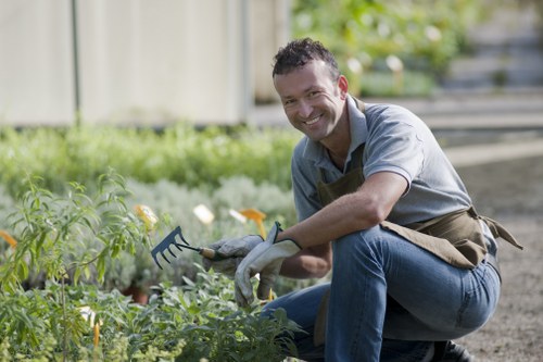 A smiling man wearing a brown apron and grey gloves is kneeling on a garden path, tending to a flower bed in a backyard garden. The flower bed contains a variety of small, leafy green plants with some yellow flowers visible. Behind him, there is a wooden fence and lush green bushes that frame the outdoor space. The garden features neatly maintained grass, a paved pathway, and a wooden deck in the background, suggesting a well-organized landscaped area. The scene occurs in natural daylight with slightly overcast weather, creating soft lighting conditions. This outdoor environment reflects a typical residential garden in Edmonton, supporting local gardening and landscaping services by showing a vibrant plant-focused outdoor space aimed at flower bed creation and garden maintenance.
